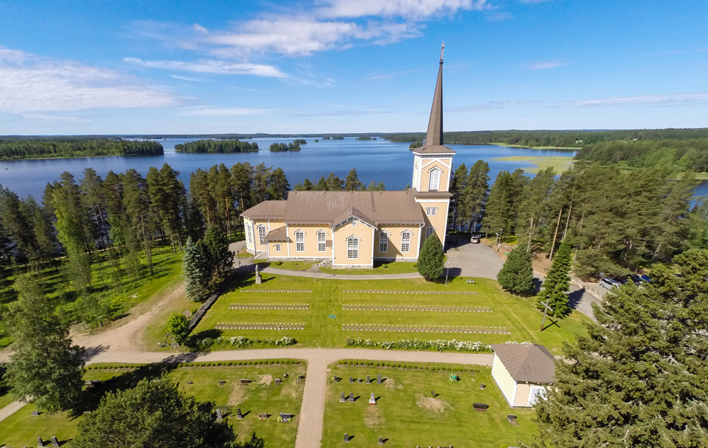 Ilmakuva Pielaveden kirkosta jossa näkyy kirkon lisäksi hautausmaata, järven rannalla suuria puita ja taust...