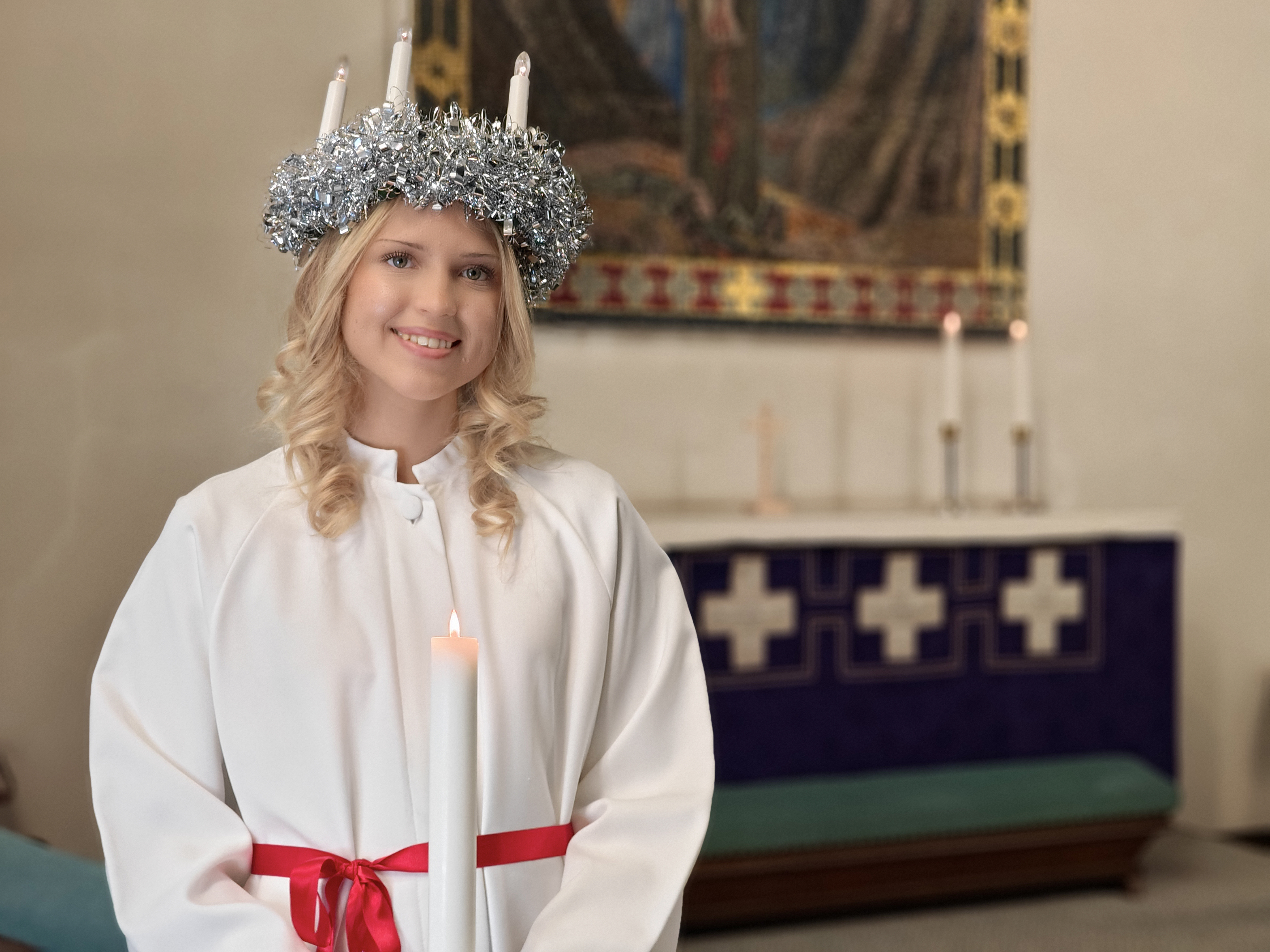 Valkopukuinen Lucia-neito kynttiläkruunu päässään ja kynttilä käsissään.