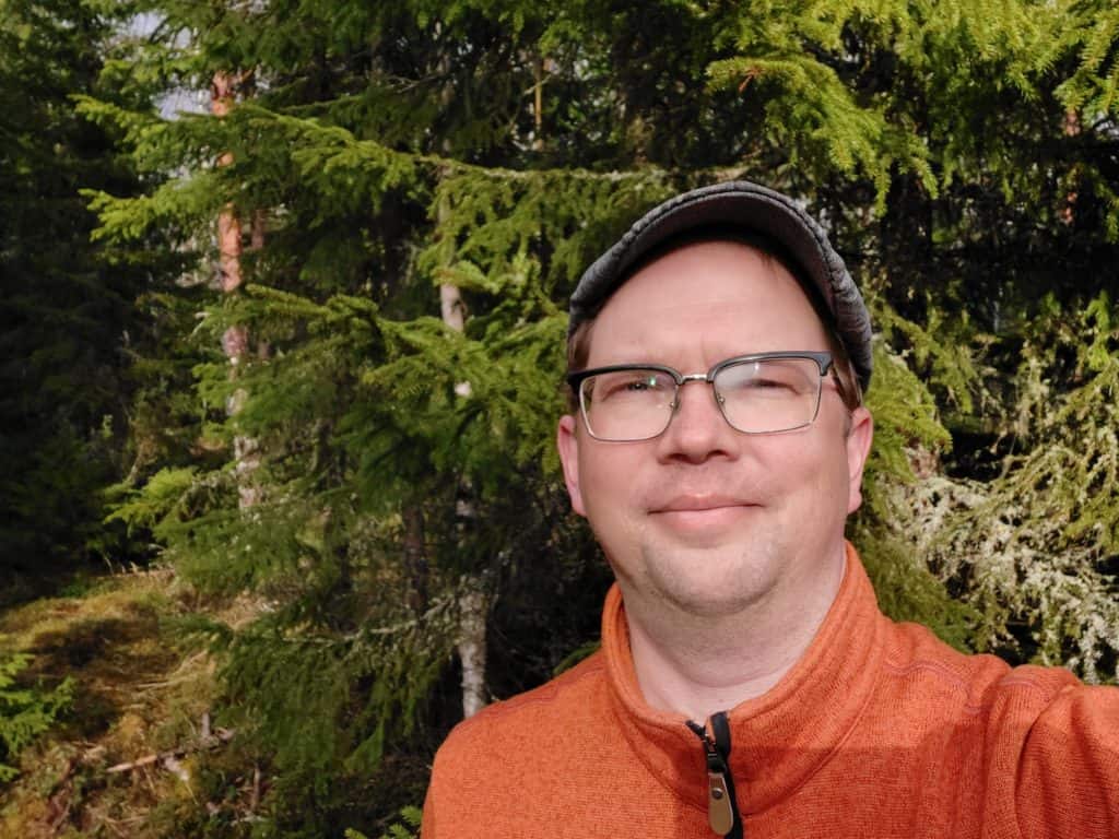 Mies hymyilee kameralle metsässä kuusikkoa vasten.