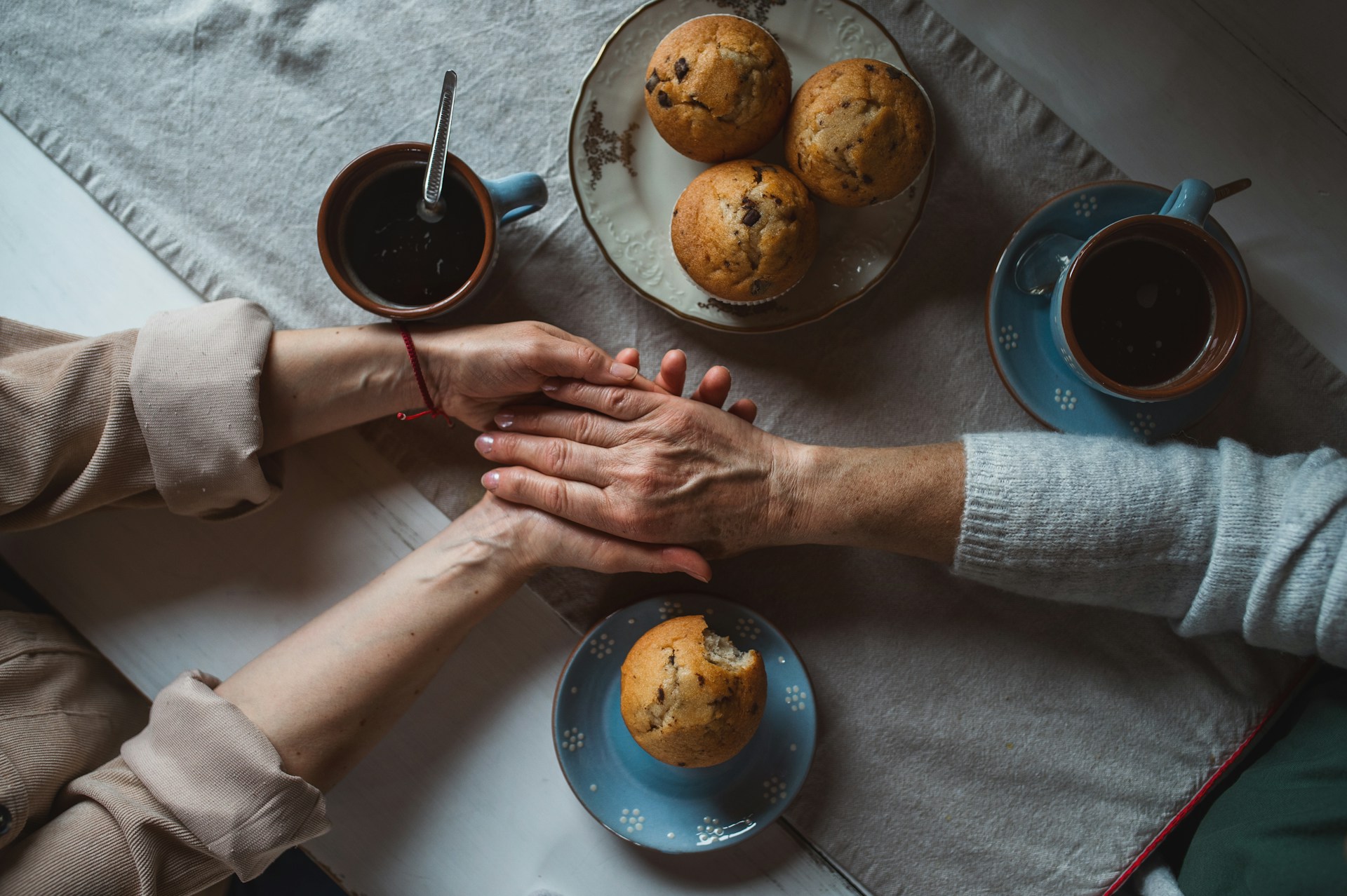 Vanhuksen ja nuoremman ihmisen kädet pöydällä, kahvikuppeja ja muffinsseja.