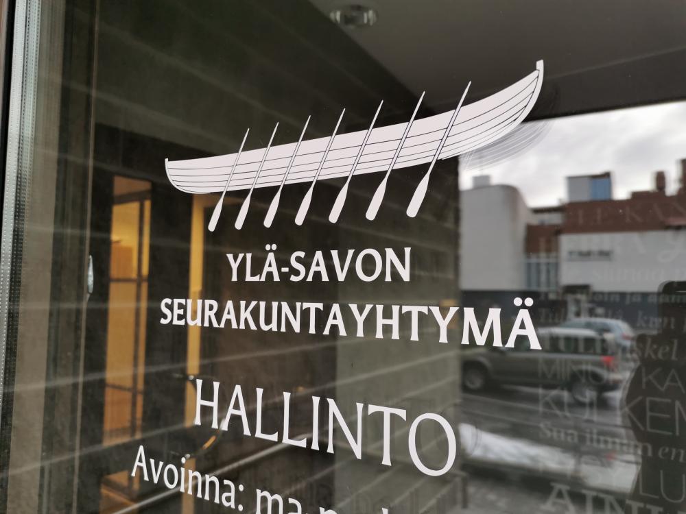 Ylä-Savon seurakuntayhtymän Hallinnon toimiston lasiovi, jossa teippaukset ja heijastuksena näkyy ympäristöä. 