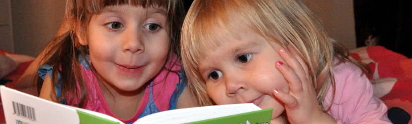 Kaksi pientä tyttöä lukemassa kirjaa silmät suurina innostuksesta, toisella käsi poskella.